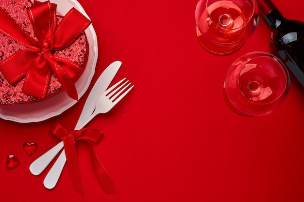 Valentijnsdag achtergrond of concept met lege witte plaat en bestek vork en mes, champagne, glazen en geschenkdoos op scharlaken of rode achtergrond. bovenaanzicht plat lag met kopieerruimte.