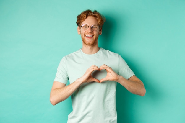 Valentijnsdag en relatieconcept. Gelukkig vriendje met rood haar en baard, bril en t-shirt dragen, hartteken tonen en zeggen dat ik van je hou, staande over turkooizen achtergrond