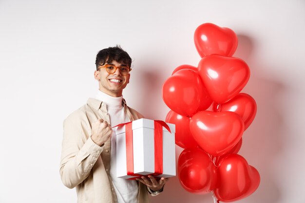 Valentijnsdag en romantiek concept. Gelukkig en zelfverzekerd vriendje bereidt cadeau voor geliefde voor, zegt ja en glimlacht, houdt een romantisch cadeau vast, staat in de buurt van rode hartenballonnen