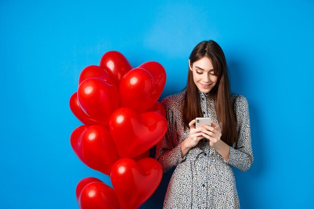 Valentijnsdag. Glimlachende vrouw in jurk staande in de buurt van rode harten ballonnen en kijken naar smartphone, staande op blauwe achtergrond.