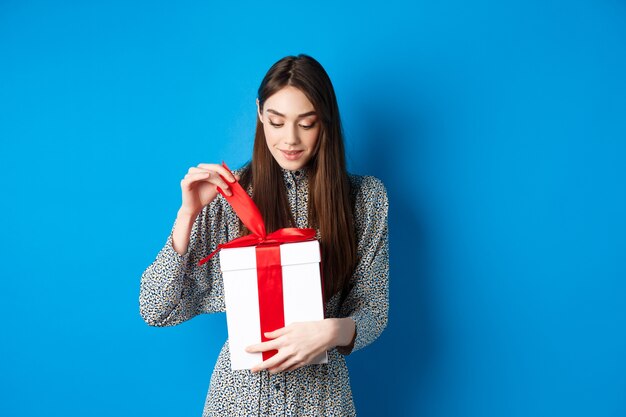 Valentijnsdag. Leuke jonge vrouw open doos met cadeau, startlint van heden en glimlachend geïntrigeerd, staande op blauwe achtergrond.