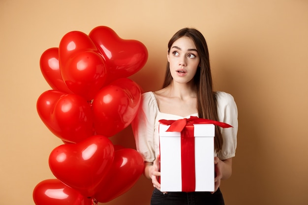 Valentijnsdag peinzend schattig meisje dat raadt wie haar cadeau heeft gedaan terwijl ze het cadeautje vasthoudt en nieuwsgierig naar je kijkt...