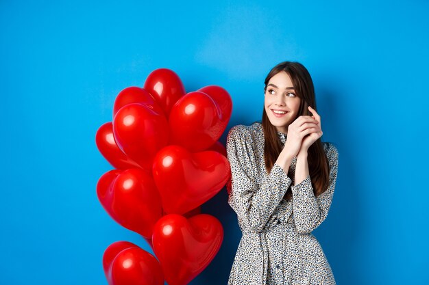 Valentijnsdag. Romantische jonge vrouw in jurk, dromerig naar links kijkend en glimlachen, staande in de buurt van rode harten ballonnen, blauwe achtergrond