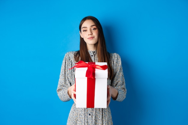 Valentijnsdag romantische vriendin brengt cadeau en glimlach naar camera in trendy jurk op blauwe b...