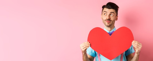 Gratis foto valentijnsdagconcept dromerige romantische man die naar links kijkt en een groot rood hartknipsel laat zien dat op staat