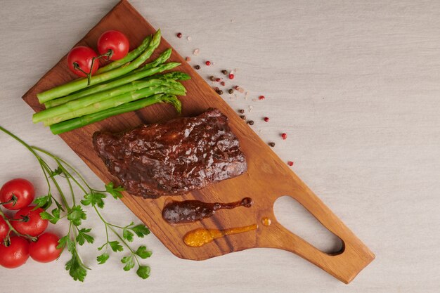 Varkensvlees geroosterde, gegrilde spareribs van een zomerse BBQ geserveerd met groenten, asperges, worteltjes, verse tomaten en kruiden. gerookte ribben op houten snijplank op stenen oppervlak. bovenaanzicht.