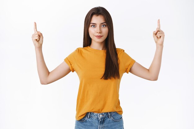 Vastberaden en assertieve aantrekkelijke zelfverzekerde brunette vrouw die rechtop staat en naar de camera kijkt met een zelfverzekerde uitdrukking die wetende blik geeft als een aanbevolen product dat op een witte achtergrond wijst