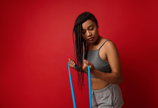 Vastberaden jonge afrikaanse vrouw in grijze sportkleding kneden biceps en schouderspieren met behulp van een elastische band van fitness tijdens een training van vetverlies, geïsoleerd op rode achtergrond met kopie advertentieruimte