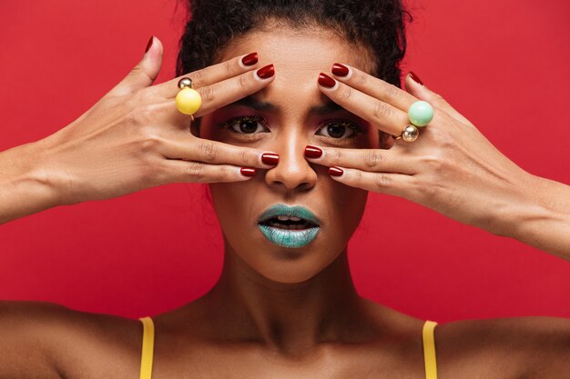 Veelkleurige jonge mulatvrouw die met trendy make-up op camera door vingers met ringen, over rode muur kijken