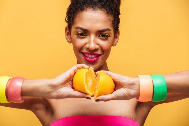 Veelkleurige joyous mulatvrouw met heldere make-up die twee delen van verse sinaasappel opnieuw samen verbindt, geïsoleerd over gele muur