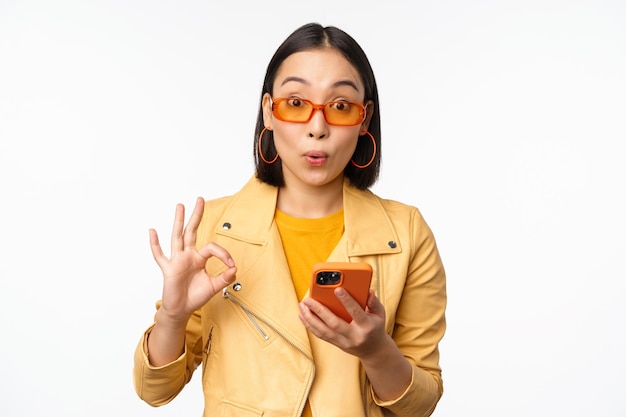 Verbaasd Aziatisch meisje met een zonnebril die een goed teken toont met een smartphone die er onder de indruk uitziet en iets aanbeveelt dat op een witte achtergrond staat