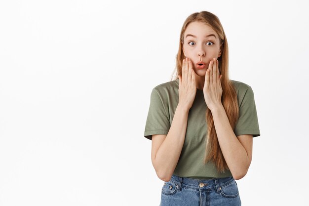 Verbaasd en onder de indruk blond meisje, hijgend gefascineerd, hand in hand bij de mond, staren vol ontzag, staande tegen de witte muur