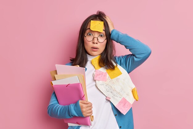 Verbaasd geschokt vrouwelijke kantoormedewerker overladen met papierwerk verbijsterd om deadline te hebben voor het afronden van onderzoek houdt mappen vast houdt hand op hoofd draagt ronde bril.