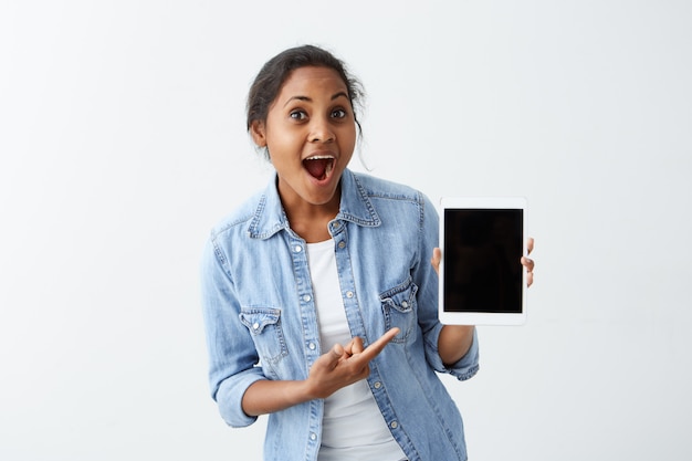Verbaasd jonge Afro-Amerikaanse jonge vrouw, gekleed in een blauw shirt over wit t-shirt met tablet in haar handen, laat zien hoe cool deze tablet is, mond wijd openhouden, verrast kijken.