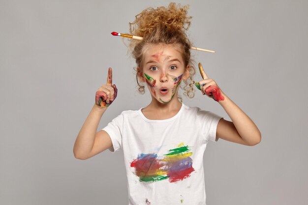 Verbaasd klein meisje met een borstel in haar chique krullende blonde haar, gekleed in een wit t-shirt, met geverfde vingers, gedraagt zich alsof ze een idee heeft, op een grijze achtergrond.