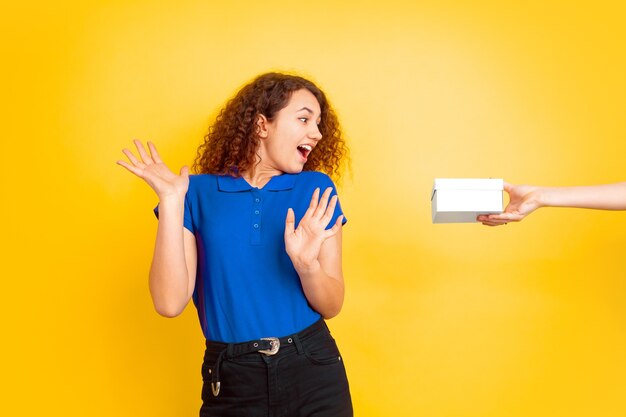 Verbaasd met het nemen van een geschenkdoos. Het meisjesportret van de Kaukasische tiener op gele muur. Mooi vrouwelijk krullend model. Concept van menselijke emoties, gezichtsuitdrukking, verkoop, advertentie, onderwijs. Copyspace.