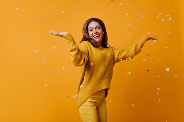 Verbaasd schattig meisje met donker haar dansen op gele muur. aantrekkelijke stijlvolle vrouw confetti weggooien.