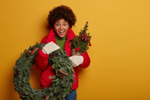 Verbaasd verbaasd Afro-Amerikaanse vrouw houdt groene krans en firtree, heeft vreugdevolle uitdrukking, bereidt zich voor op Kerstmis of Nieuwjaar, geïsoleerd over gele muur.