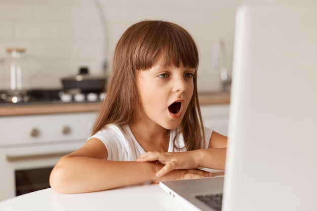 Verbaasd verbaasd donkerharig vrouwelijk kind zittend aan tafel, kijkend naar laptopscherm met wijd open mond, ziet iets verbazingwekkends, poserend in de keuken.