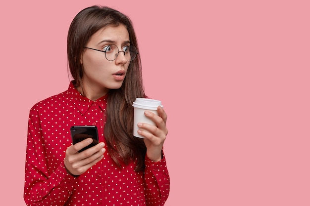 verbaasd verrast donkerharige mooie vrouw houdt afhaalmaaltijden koffie, moderne mobiele telefoon, gekleed in rode blouse