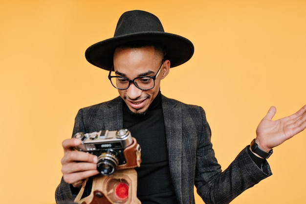 Verbaasd zwarte man in hoed camera kijken. portret van emotionele Afrikaanse mannelijke fotograaf die zich op gele muur bevindt.