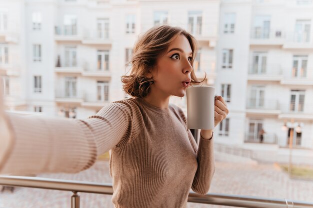 Verbaasde blanke dame in stijlvolle trui genieten van thee. Charmant krullend vrouwelijk model kopje koffie houden en selfie maken op balkon.