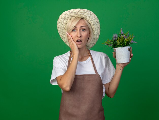 Verbaasde blonde tuinmanvrouw van middelbare leeftijd in uniform met een hoed die een bloempot vasthoudt en haar hand op het gezicht houdt