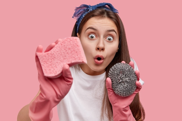 Verbaasde brunette dame met schoonmaakgereedschap, ogen uitgesprongen, geschokt om veel vuil te zien