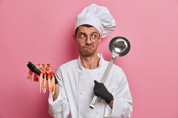 Verbaasde chef-kok houdt rauwe rivierkreeft en pollepel vast, gaat soep bereiden van zeevruchten, draagt uniform, hoed, ronde bril, kookt diner in restaurant
