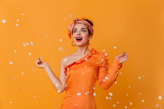 Verbaasde dame in oranje jurk met blote schouder en Africanstyle hoofdband poseren op oranje achtergrond onder confetti