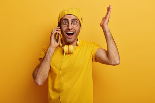 Verbaasde emotionele man steekt zijn hand op, reageert op schokkende relevantie, heeft telefoongesprekken met iemand, draagt een stijlvolle gele hoed