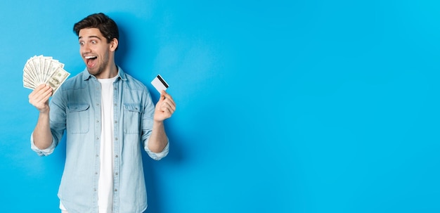 Gratis foto verbaasde en gelukkige man met creditcard kijkend naar geld tevreden staande over blauwe achtergrond