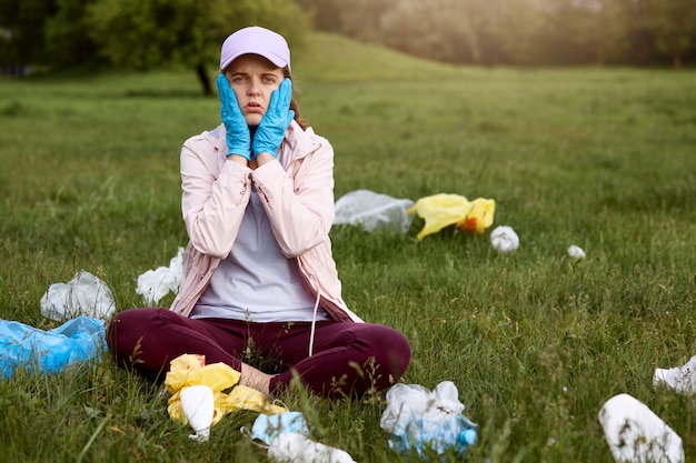 Gratis foto verbaasde en geschokte vrouw die in het park op groen gras zit met palmen op de wangen, nonchalant gekleed, omringd door afval, moet al het afval oppikken.