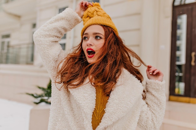 Verbaasde gembervrouw die haar gebreide muts aanraakt Foto van een emotioneel blank meisje dat in de winter poseert