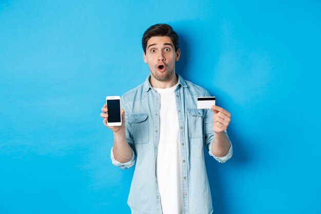 Verbaasde jonge man die het scherm van de mobiele telefoon en creditcard toont, online winkelt, staande tegen een blauwe achtergrond