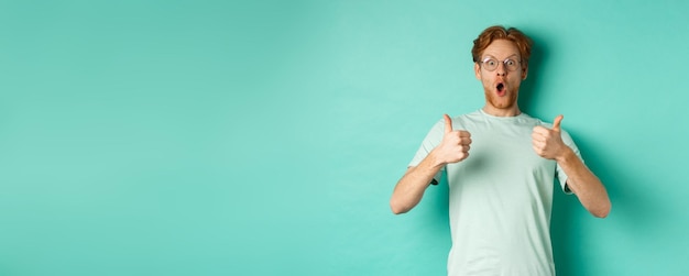 Gratis foto verbaasde jonge man met rood haar en baard die een bril draagt met een t-shirt dat duim omhoog laat zien en hijgend naar binnen