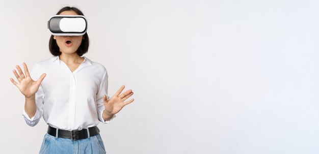 Verbaasde jonge vrouw in virtual reality die een vr-brilhoofdtelefoon gebruikt die over een witte achtergrond staat