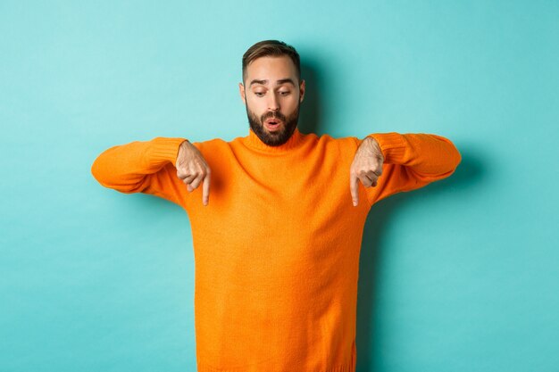Verbaasde man in oranje trui, wijzend met zijn vingers naar beneden en kijkt geïnteresseerd over de turquoise muur.