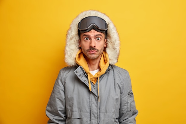 Verbaasde man snowboarder staart afgeluisterde ogen draagt warme winterjas met capuchon rust tijdens koude december kan niet geloven in iets ongelooflijks.