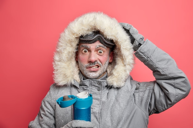 Gratis foto verbaasde mannelijke wandelaar heeft bevroren gezicht geschokt door koude omstandigheden tijdens expeditie draagt warme jas en skibril drinkt warme drank.