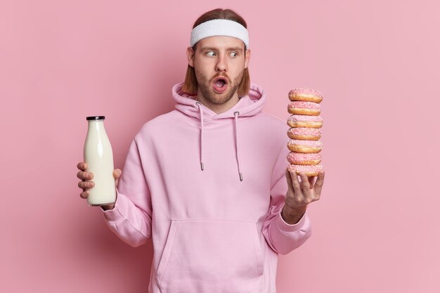 Verbaasde sportieve man staart naar smakelijke heerlijke donuts heeft de verleiding om te eten gekleed in een witte hoofdband met capuchon.