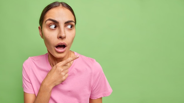 Verbaasde verbaasde donkerharige vrouw gekleed in casual t-shirt geeft weg op lege kopie ruimte houdt mond open en reageert op verbazingwekkende aanbieding geïsoleerd over groene achtergrond. Omg kijk daar.