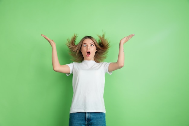 Verbaasde, verbaasde jonge vrouw die op groene studiomuur wordt geïsoleerd