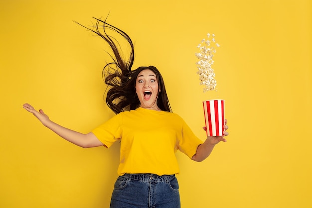 Verbaasde, vliegende popcorn. Blanke vrouw portret geïsoleerd op gele muur. Mooi donkerbruin model in casual. Concept van menselijke emoties, gezichtsuitdrukking, verkoop, advertentie, copyspace.