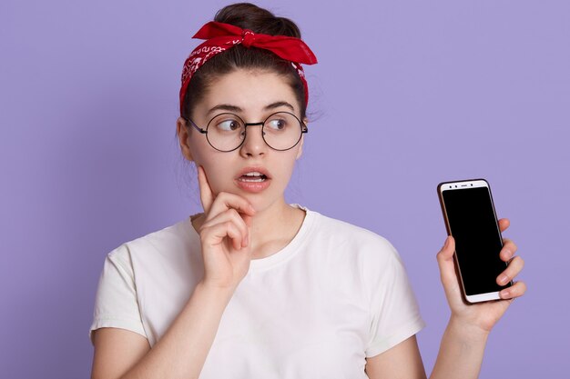 Verbaasde vrouw met vinger op wang, kijkt opzij met geopende mond en verbaasde gezichtsuitdrukking, dame in wit casual t-shirt en rode haarband