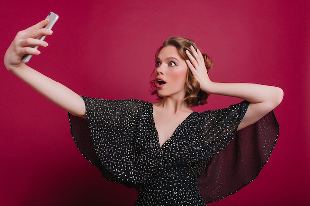 Verbaasde vrouw speelt met kort glanzend haar tijdens het maken van selfie. Geschokt Kaukasisch meisje in vintage casual kleding foto van zichzelf, met behulp van smartphone.