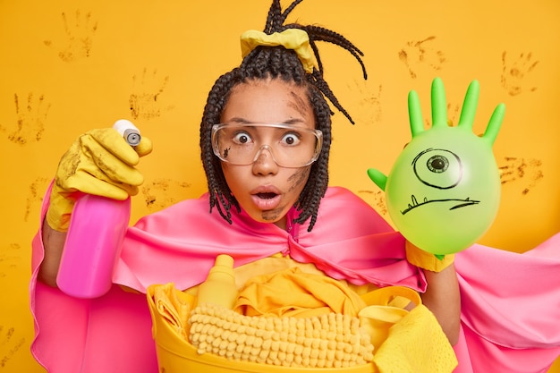 Verbaasde vrouwelijke huishoudster met donkere huid staart naar camera houdt wasmiddel vast en opgeblazen ballon draagt grote transparante glazen cape doet alsof ze een superheld is die klaar is om schoon te maken of vuil te verwijderen