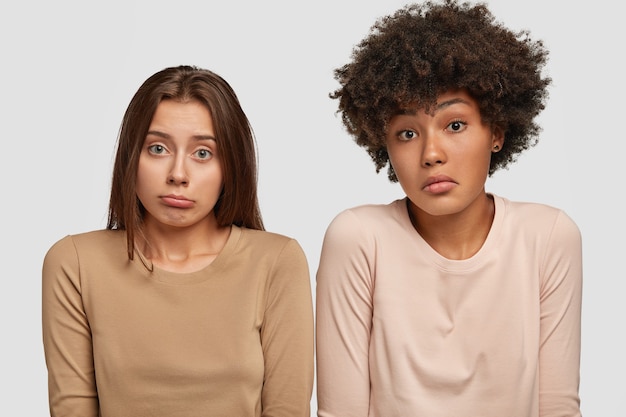 Gratis foto verbaasde vrouwen halen hun schouders op in verbijstering: mooie europese vrouw staat dicht bij afro-amerikaanse vriendin, kan geen beslissing over iets nemen, geïsoleerd over witte muur.