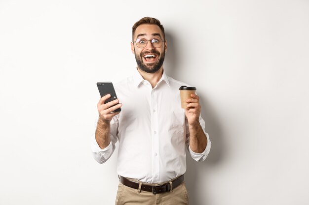 Verbaasde zakenman die koffie drinkt, reageert op geweldig online aanbod op mobiele telefoon, staand