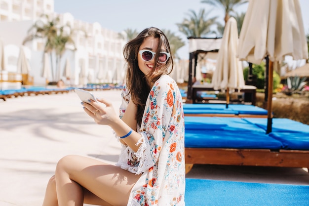 Verbazend gelooid meisje met donker haar lachen, zittend op een blauw schraagbed bij het zwembad. prachtige brunette jonge dame in een stijlvol shirt met bloemen versiering met haar telefoon en lacht naar iemand